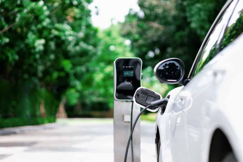 Electric vehicle charging point connected to a white car on a green outdoor background.