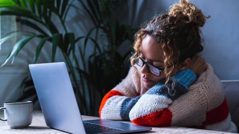 Overwhelmed woman with glasses resting her head on her arms at a laptop in a cozy home office.