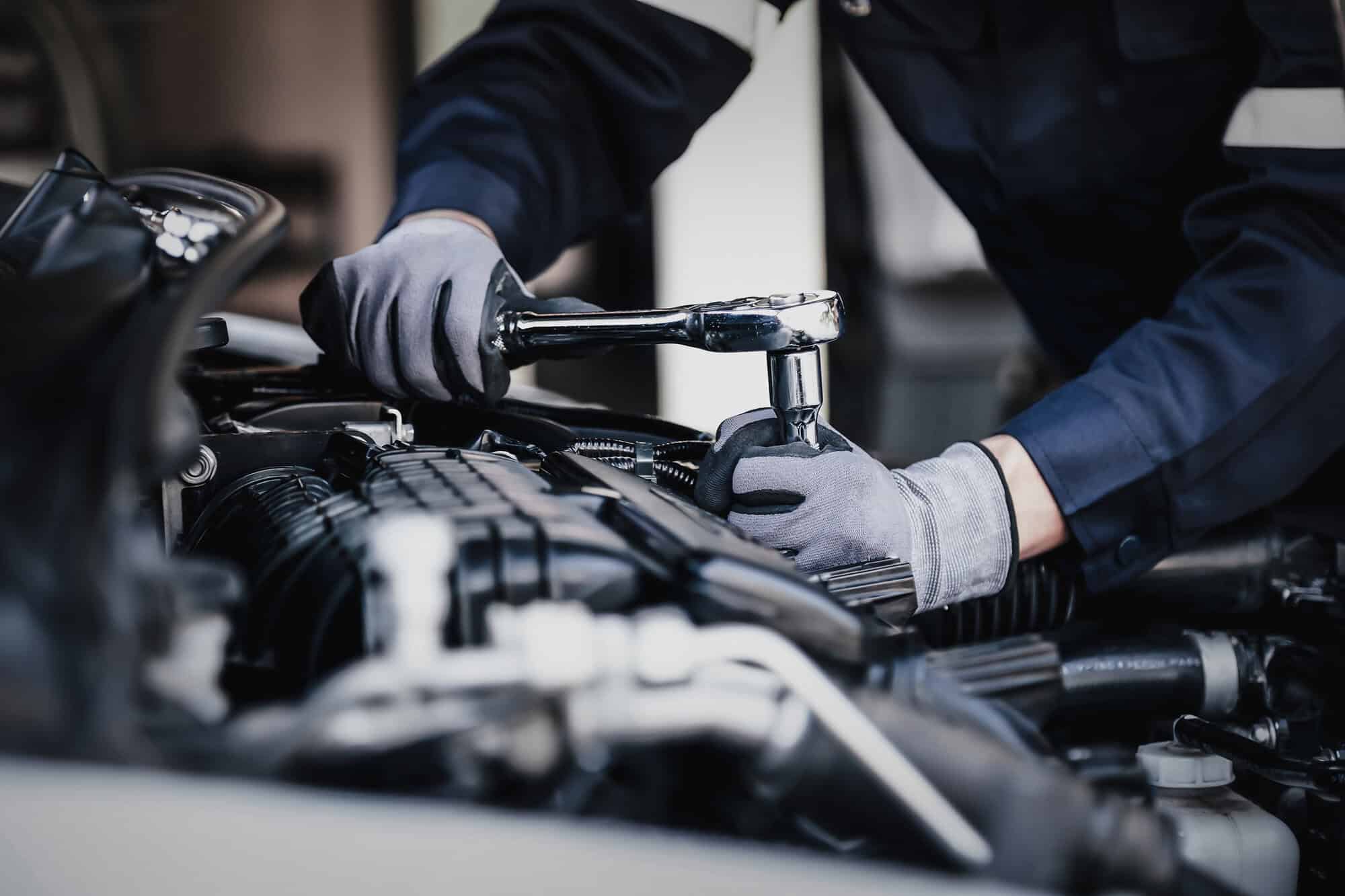 Close-up of a vehicle engine being serviced in a garage, with gloved hands using a ratchet wrench to carry out maintenance work. The engine components are in focus, highlighting professional motor trade repair and servicing activities.