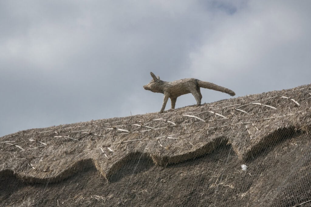 A close-up of a thatched roof protected with wire mesh, featuring a decorative fox figure made from thatch standing along the roof ridge beneath a cloudy sky.