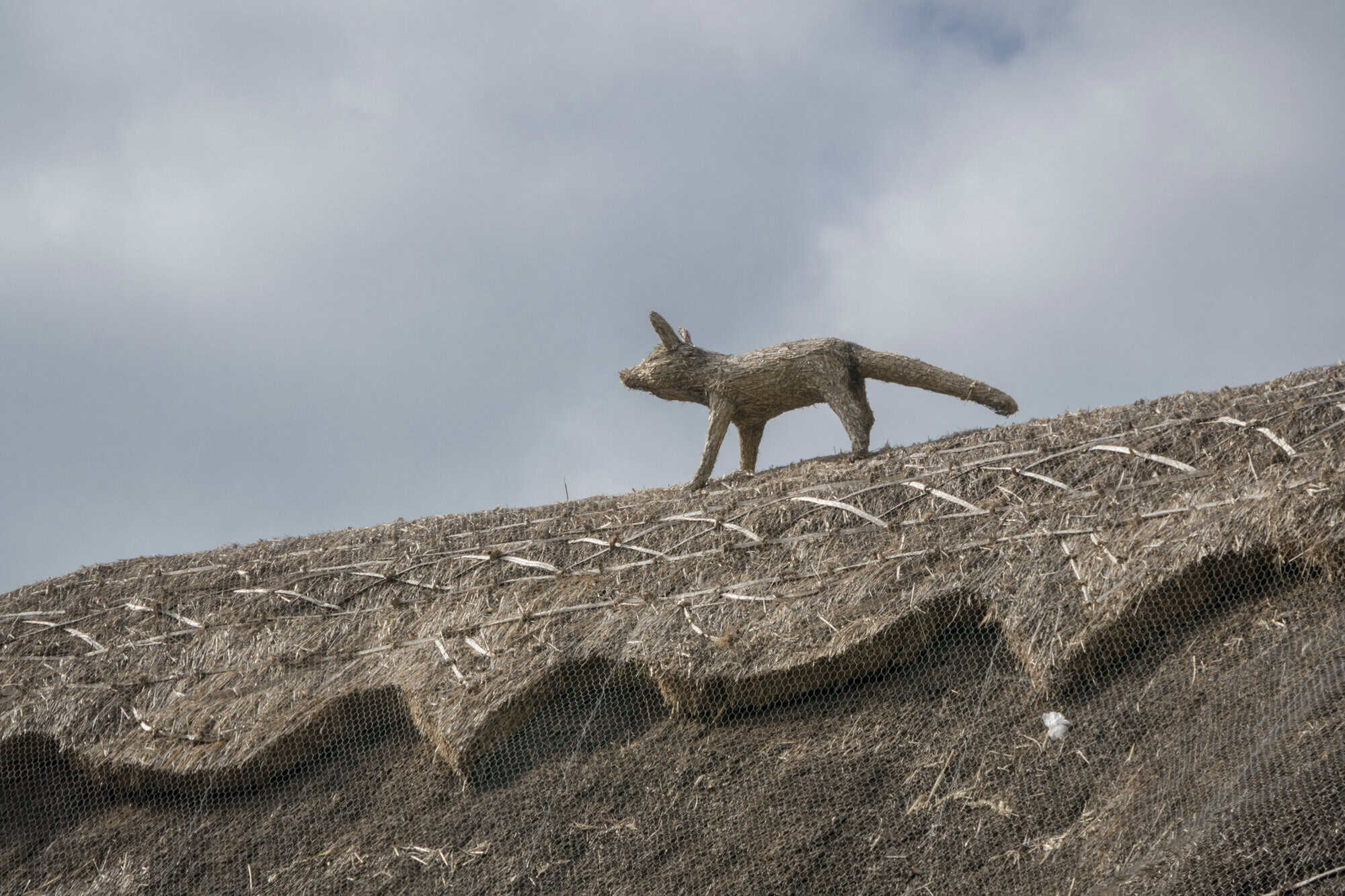 A close-up of a thatched roof protected with wire mesh, featuring a decorative fox figure made from thatch standing along the roof ridge beneath a cloudy sky.