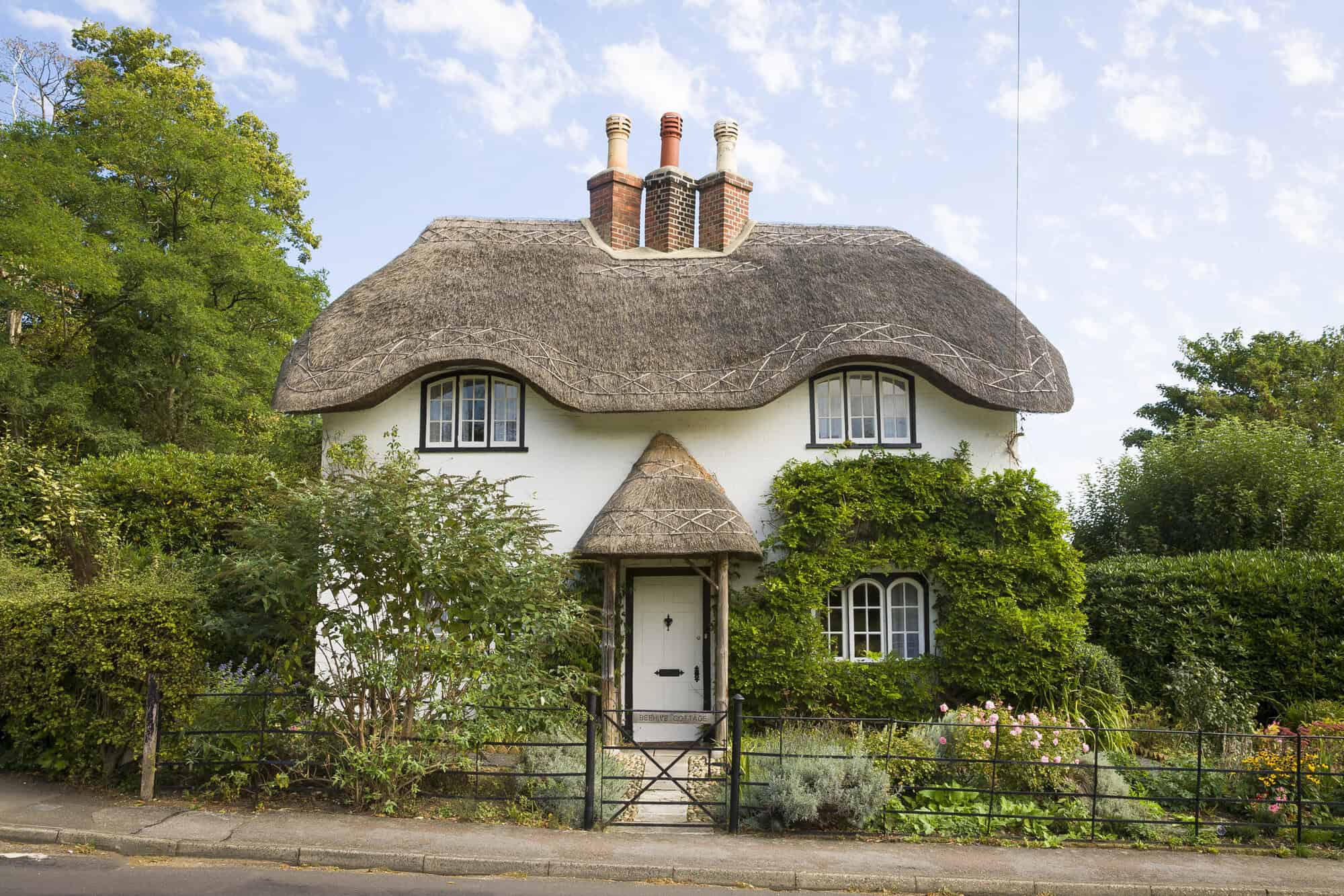 A traditional white thatched cottage with a curved straw roof, three brick chimneys, and a small thatched porch, surrounded by a well-maintained garden and greenery under a bright blue sky.