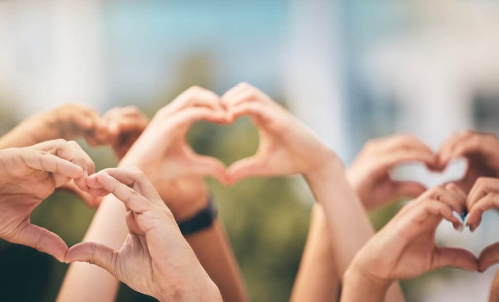 Multiple hands in the foreground form heart shapes against a softly blurred outdoor background. The image symbolises community, support, care, generosity and charitable giving, with natural light creating a warm and welcoming atmosphere.