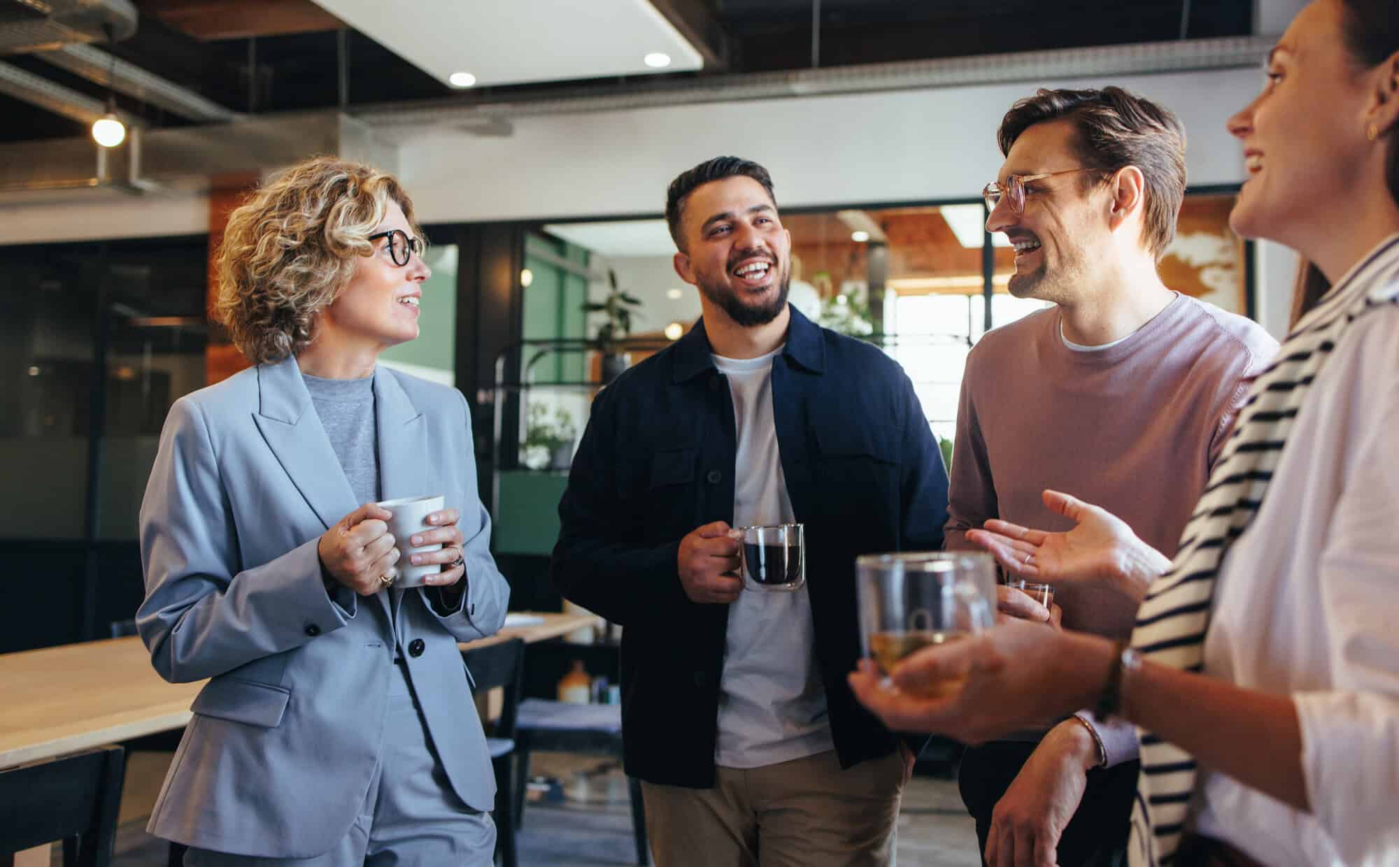 Group of colleagues standing together in a modern office, holding drinks and chatting during an informal workplace meeting or break. The open-plan office features glass partitions, natural lighting, and collaborative workspaces in the background.
