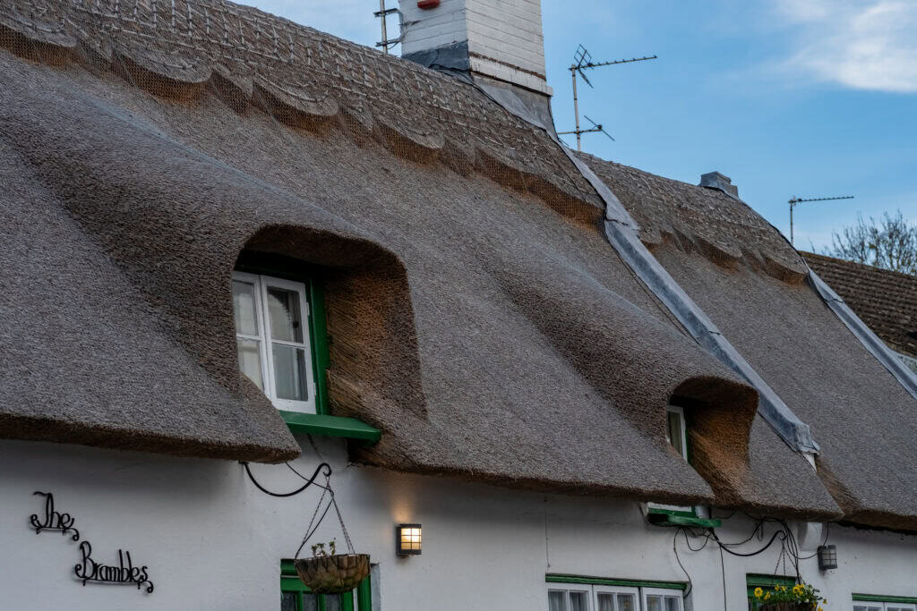 Close-up of a traditional whitewashed thatched cottage with a thick straw roof, dormer windows with green trim, and a chimney. Protective wire netting is visible across sections of the thatch, with exterior lights and hanging baskets mounted on the front of the property beneath a blue sky.